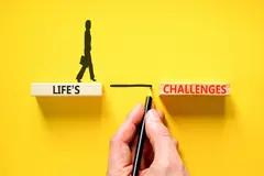 Hand drawing a line between two wooden blocks on a yellow background marked with the words LIFE'S and CHALLENGES enabling a person to cross the gap