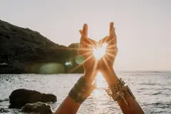 Woman's two hands making a heart shape with the sun coming through the gap. She's wearing boho style braclets practicing yoga outdoors by sea on sunset
